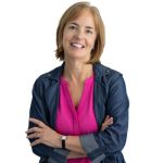 A woman, Gail Slater, Vice President of Development & Communication is standing against a plain white background, smiling warmly at the camera. She has short, light brown hair and is wearing a bright pink blouse under a denim jacket. Her arms are comfortably crossed, and she appears relaxed and approachable. The clean background and casual-professional attire give the portrait a friendly, modern feel.