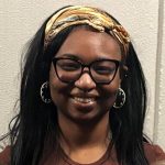 A headshot of Kharisma Thomas a black woman wearing a gold head band, black glasses and silver earrings, smiling at the camear.